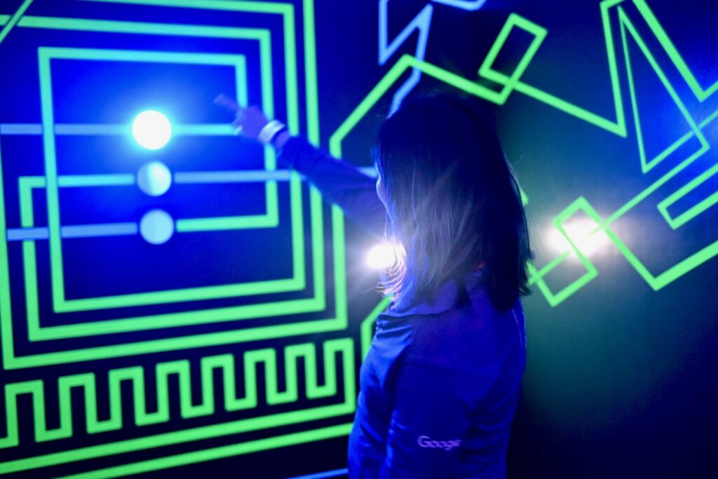 A dark room with glowing green patterns on the wall. A woman points at a bright light.