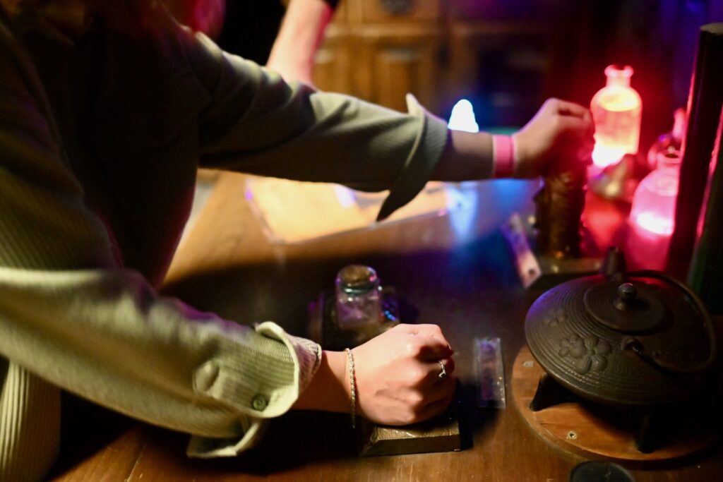 A table with glowing bottles and an old kettle. A person touches a different bottle with each hand.