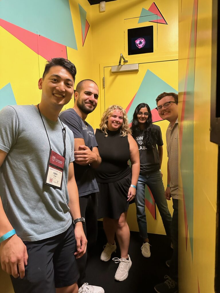 Five people smile for a photo in a yellow room with colored triangles on the walls.