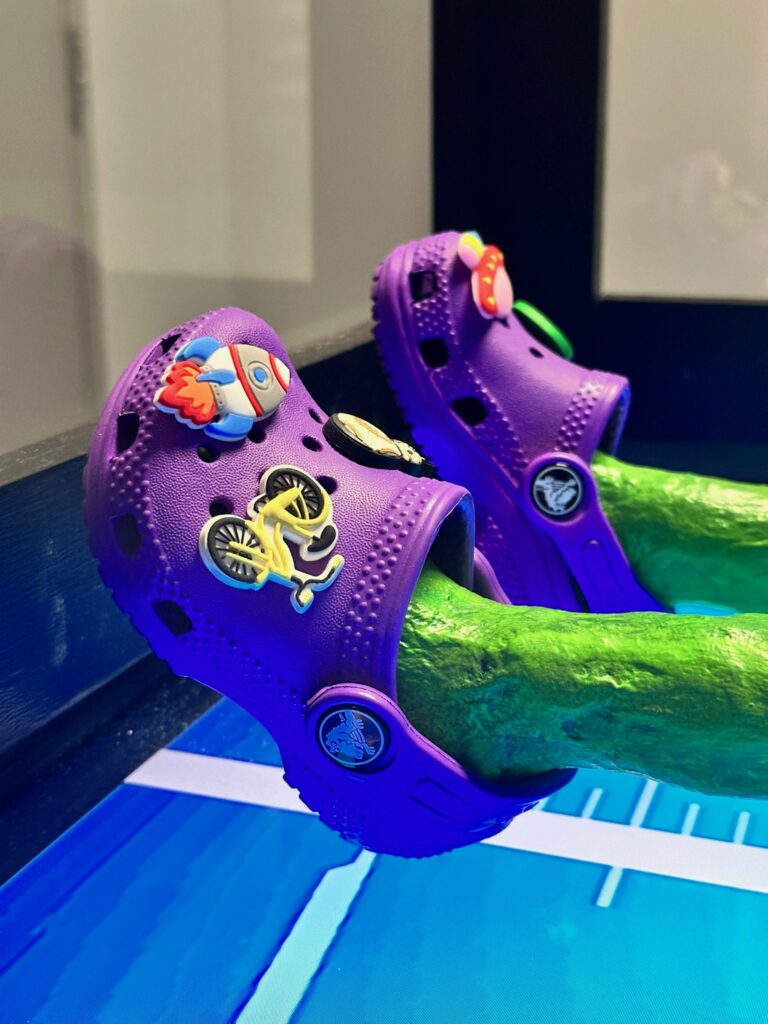 Thin green legs wearing purple crocs with a rocket charm and a bicycle charm.