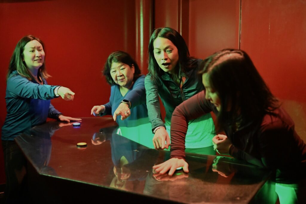 Four women at a control table. One woman presses a button while the others point at it with excited urgency.