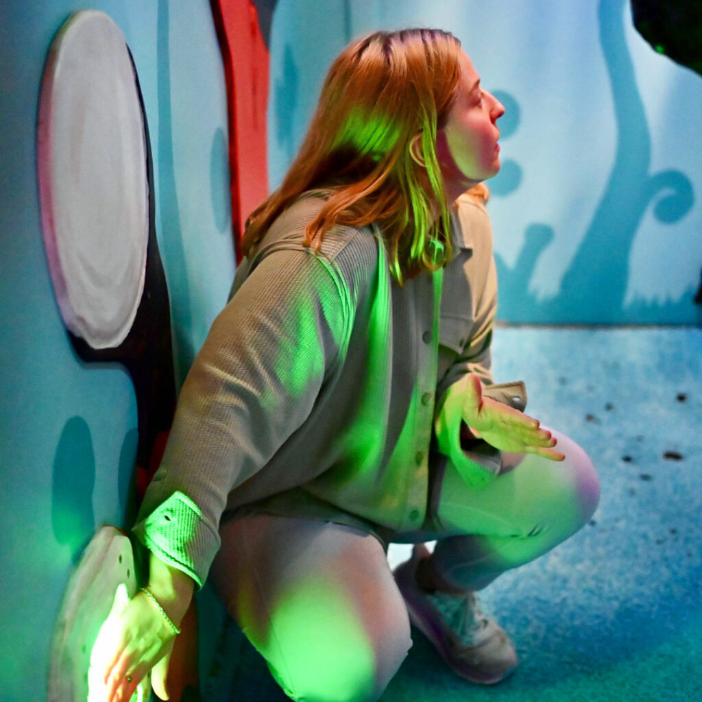 A woman in a surreally painted room crouches to push a glowing panel.