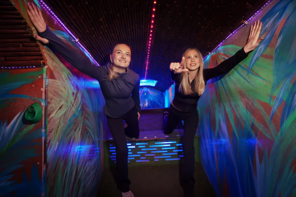 två glada tjejer kliver ned för en smal korridor med lutande golv, den ena pekar framför sig